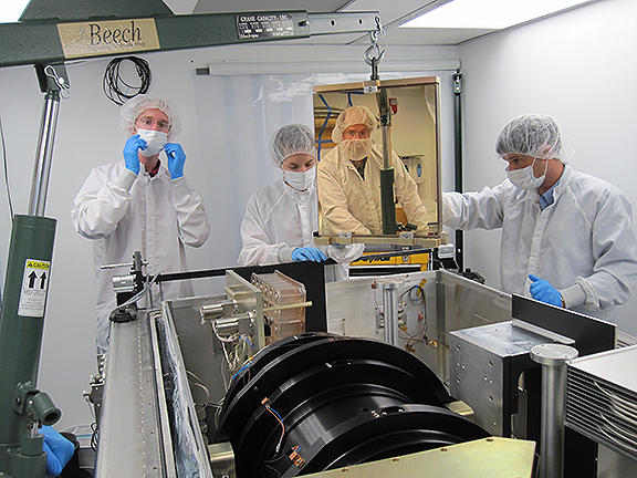 Four people in white clean suits and masks stand around the APOGEE spectrograph, a black cylinder about a meter long